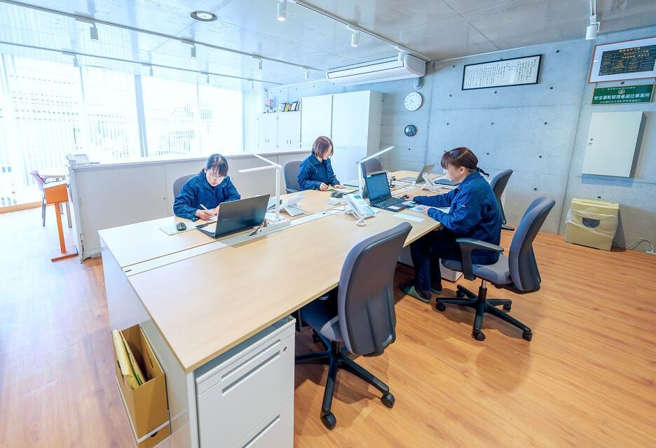 株式会社ヒタチ設備のオフィスで、デスクに向かいノートパソコンで業務を行う作業着姿の社員。