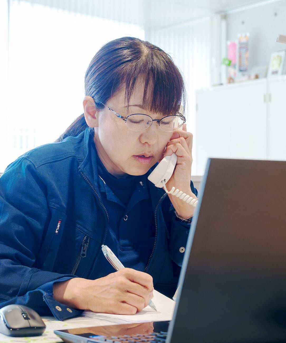 株式会社ヒタチ設備の社員インタビュー。オフィスで電話応対をしながら事務作業を行う、作業着姿の女性社員。