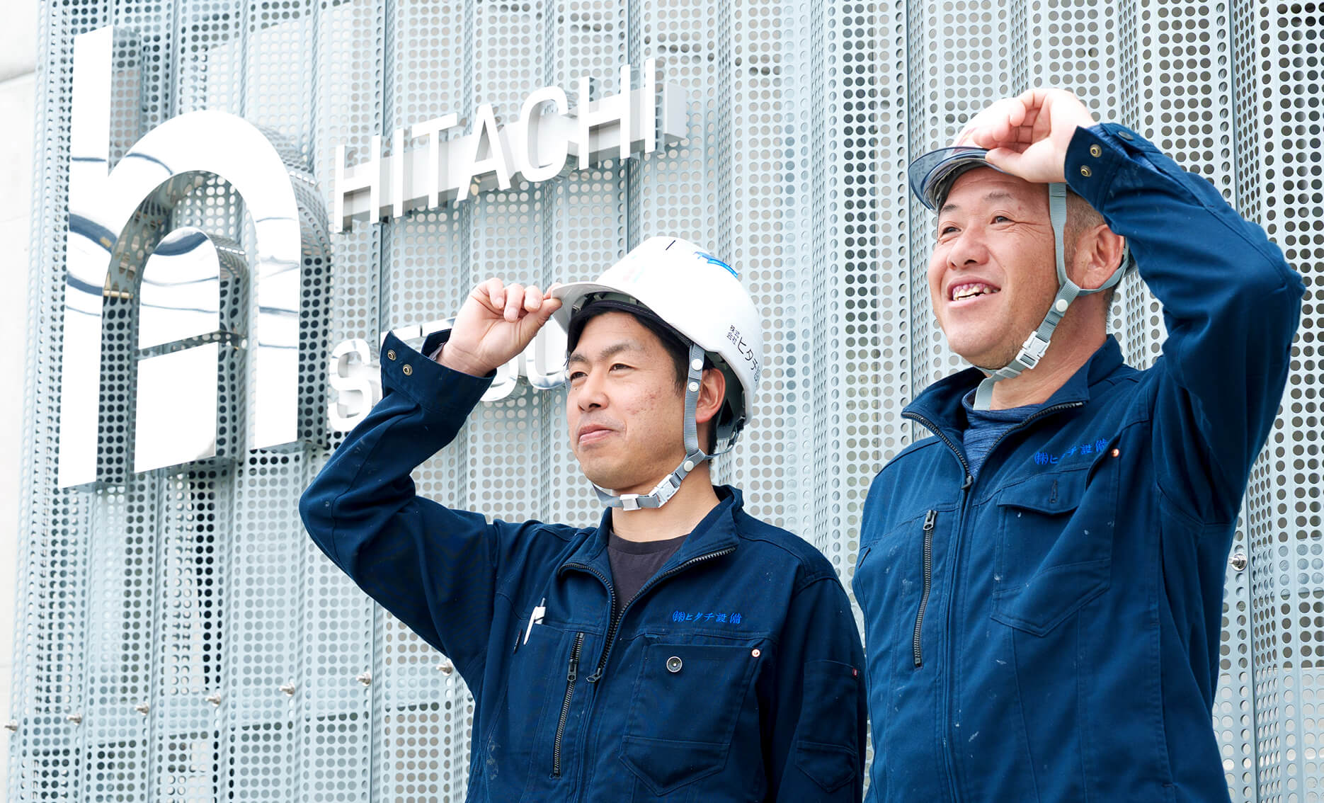 株式会社ヒタチ設備の社屋前で笑顔を見せる、ヘルメットと作業服を着用した2人のスタッフ