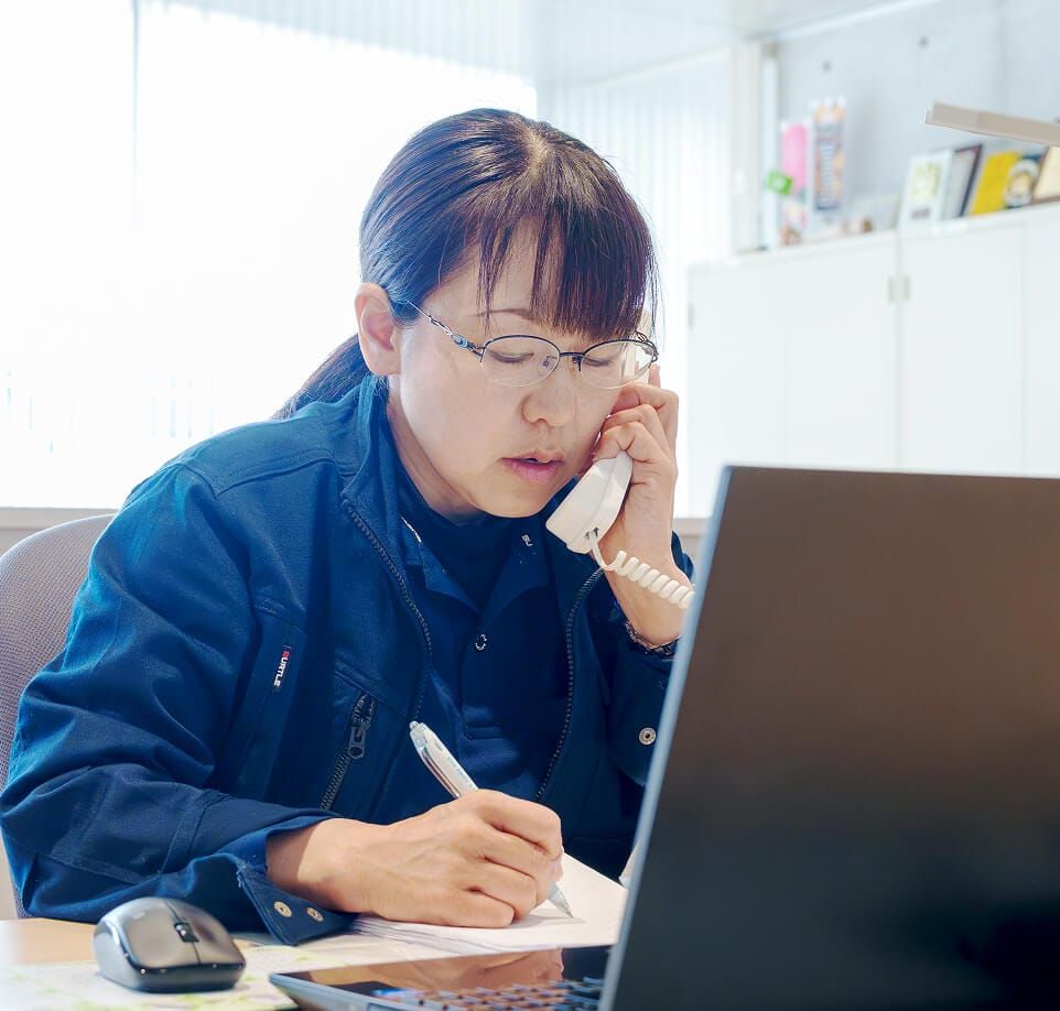 オフィスで電話対応をしながら、パソコンの前でメモを取る作業服姿の女性従業員