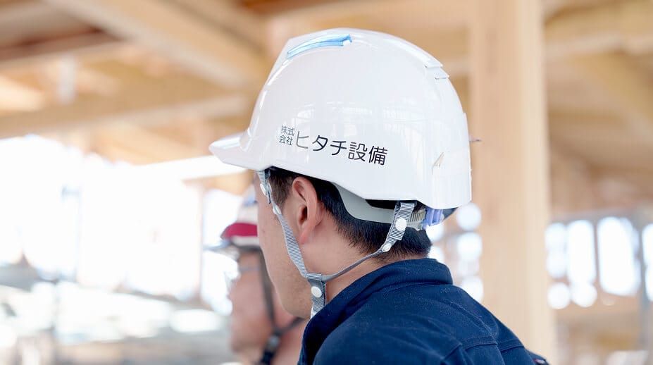 「株式会社ヒタチ設備」のロゴ入りヘルメットを被り、施工現場に立つスタッフの姿