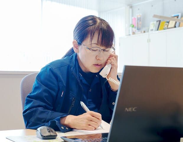 ヒタチ設備のオフィスで電話対応をしながらメモを取る事務スタッフの様子