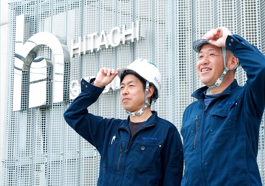 ヒタチ設備のロゴの前で作業服を着用し、笑顔で外を見つめる2人のスタッフ。